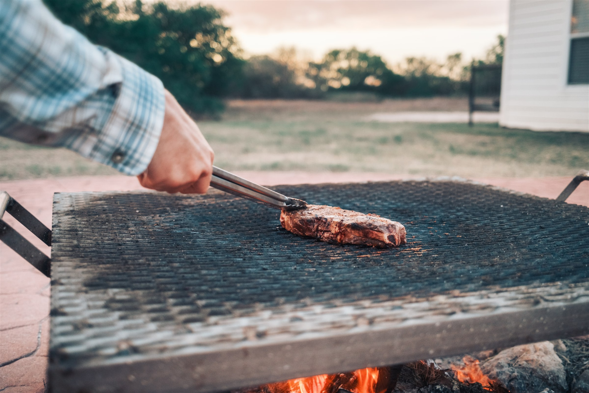 A hand turning a thick Sussex Ranch steak with tongs over an open flame at sunset, with pasture in the background